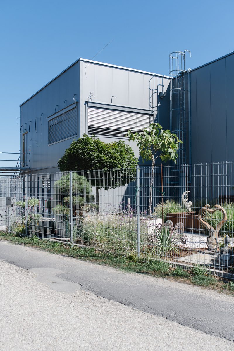 Fertiggestelltes Gebäude mit dunkler Fassadenverkleidung hinter einem Zaun mit begrüntem Vorgarten