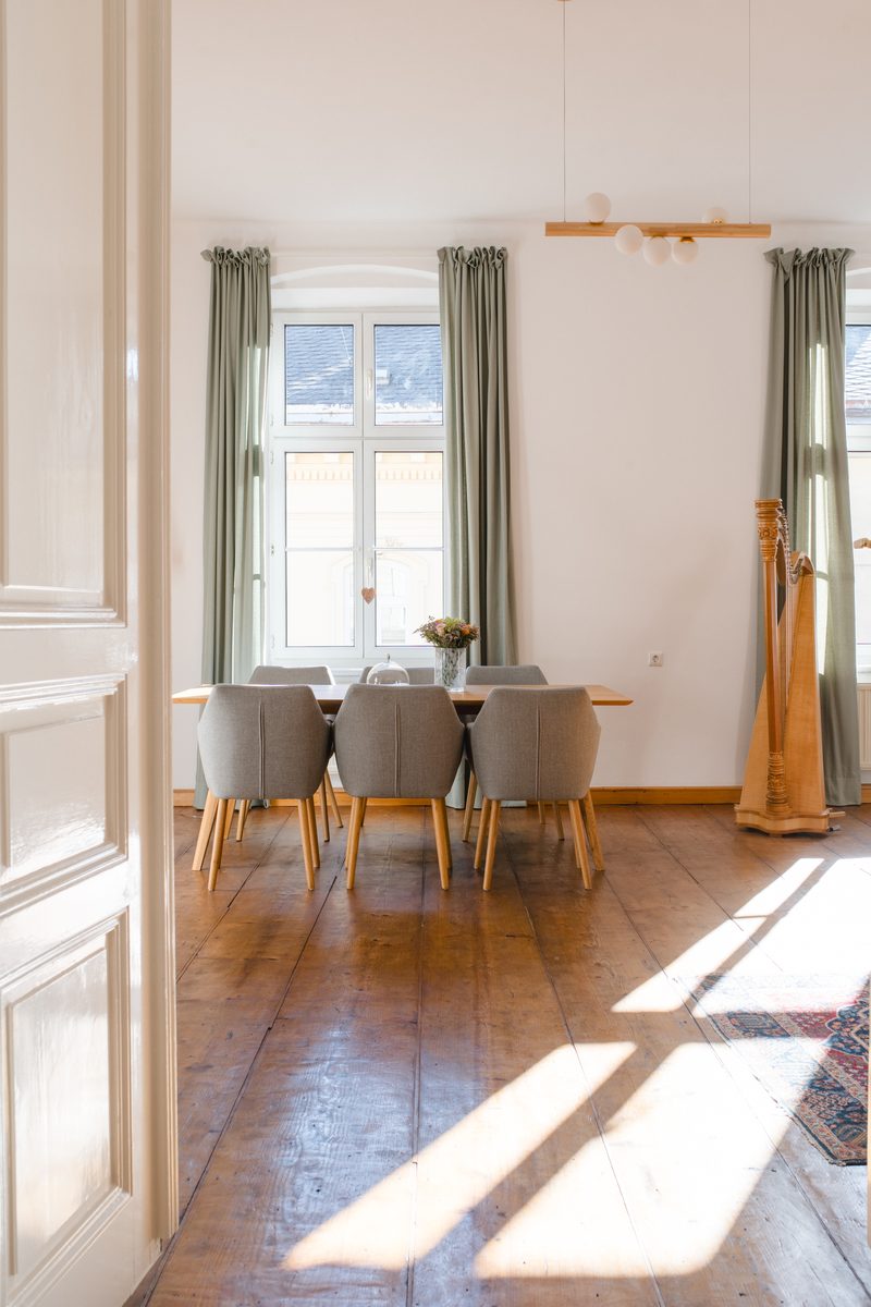 Esszimmer mit gedecktem Tisch, gepolsterten Stühlen und einer Harfe vor hohen Fenstern mit grünen Vorhängen